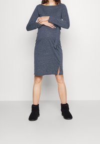 Těhotná žena oblečená v dlouhých modrých šatech a černých kotníkových botách, stojící s rukama složenýma přes břicho před jednobarevným pozadím.