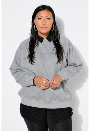 Šedá oversized mikina s žebrovanými manžetami, vrstvená přes černou košili s proužky. Model předvádí ležérní styl s doplňky.