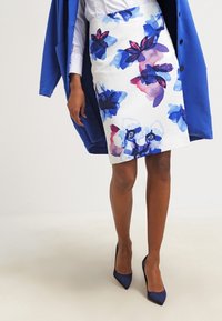 Bleistiftrock mit Blumenmuster in Weiß mit blau-rosa Designs, strukturierter Stoff, kombiniert mit einem blauen Blazer und passenden High Heels.