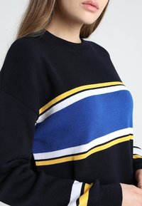 Czarny sweter z poziomymi paskami w kolorze niebieskim, białym i żółtym. Żebrowany dekolt i mankiety, w luźnym kroju. Miękka dzianina.