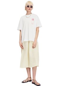 Witte oversized t-shirt met rode graphic, gecombineerd met een gele gestreepte midi rok. Accessoires zijn een meerkleurige kralenketting en een zonnebril.