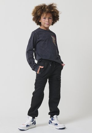 Un niño con cabello rizado está de pie con las manos en los bolsillos, vistiendo una camisa negra de manga larga, pantalones negros y zapatillas blancas y azules sobre un fondo blanco.