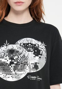 Melna T-kreklu ar lielu sudraba diskotēkas bumbas druku un baltu tekstu "Disco Fever Night Party" zem tā. Apaļš kakla izgriezums un īsas piedurknes.