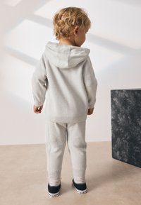 Sudadera con capucha gris claro con textura en forma de chevron, pantalones a juego y zapatillas negras; niño con el conjunto de espaldas a la cámara.