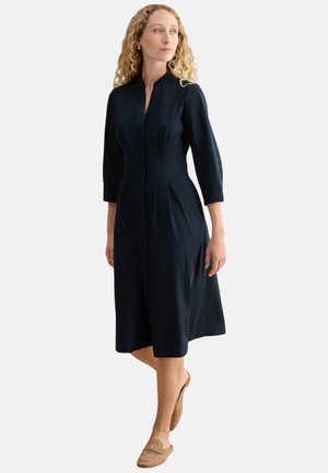 Frau mit lockigem blondem Haar, die ein dunkles, knielanges Kleid mit dreiviertel Ärmeln und beigen Slippern trägt, steht auf weißem Hintergrund.