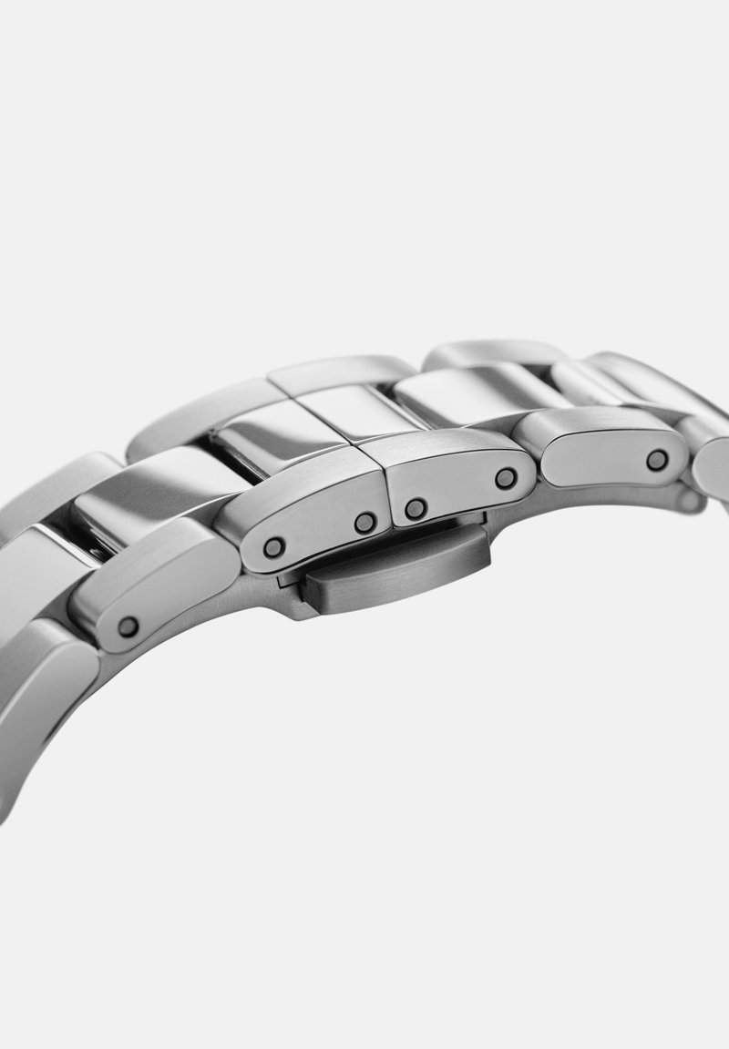 Bracelet de montre en acier inoxydable avec maillons polis, présentant des bords arrondis, des rivets noirs et un mécanisme de fermoir sécurisé.