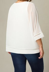 Donna che indossa una camicetta bianca trasparente con bordo in pizzo su maniche e orlo, abbinata a jeans blu scuro, vista da dietro.