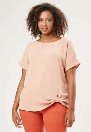 Perzikkleurige blouse met korte mouwen en een ronde halslijn, met een geknoopt accentdetail aan de onderkant, gecombineerd met oranje broeken.