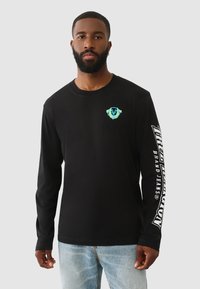 Schwarzes Langarmshirt aus Baumwolle. Mit einem kleinen türkisfarbenen Grafikdesign auf der linken Brust und weißem Text entlang des rechten Ärmels.