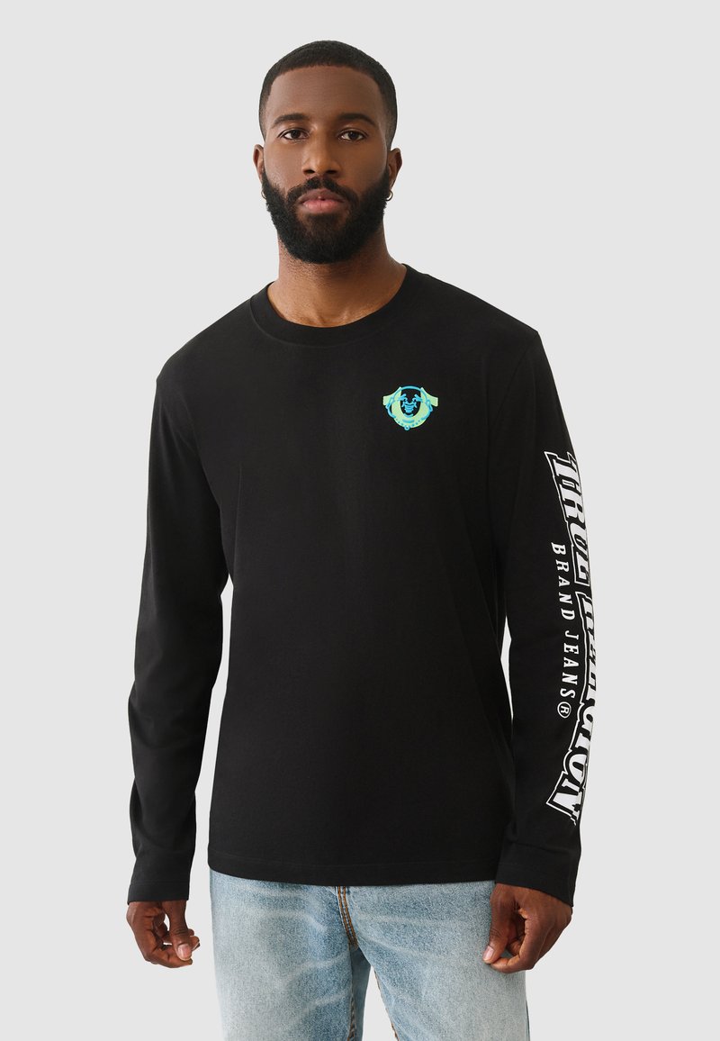 Schwarzes Langarmshirt aus Baumwolle. Mit einem kleinen türkisfarbenen Grafikdesign auf der linken Brust und weißem Text entlang des rechten Ärmels.