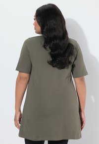 Frau mit langen schwarzen Haaren, die eine olivgrüne Tunika mit kurzen Ärmeln trägt, steht und ist von hinten vor einem schlichten weißen Hintergrund zu sehen.