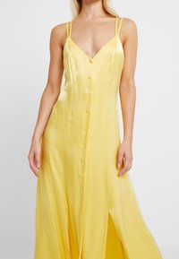 Robes maxi en satin jaune avec fines bretelles, décolleté en V profond et boutons à l'avant. Présente des fentes latérales pour plus de mouvement et de fluidité.