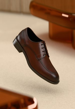 Zapato de vestir de cuero marrón pulido con cordones negros, suela negra y tacón, exhibido sobre una superficie texturizada beige y muebles de madera.