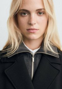 Abrigo de lana negro con un cuello estructurado, superpuesto a una camiseta de punto gris con cierre de cremallera y un cuello interior blanco. Cabello rubio claro.