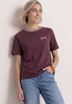 Lyshåret kvinde smiler, iført en lilla t-shirt med korte ærmer og et lille sløjfedesign, lyseblå jeans og et ravarmbånd, står mod en lys væg.