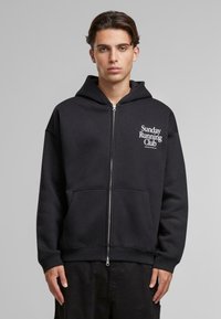 Felpa nera con zip e tascone con una stampa grafica "Sunday Running Club" sul lato sinistro superiore. Tessuto morbido, vestibilità rilassata.