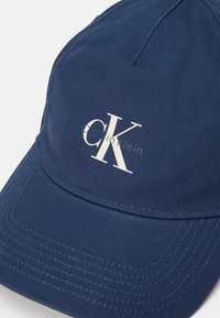 Navy baseball cap made of cotton with a curved brim, featuring white "CK" logo and stitching details on the brim and crown area.