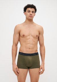 Boxer-briefs pour hommes en tissu vert olive avec une ceinture élastique bleue portant le texte "ARMOUR". Coutures latérales et design de poche à l'avant.