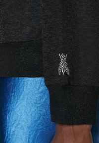 Pull noir avec une finition texturée et un poignet côtelé, arborant un accent en strass ressemblant à une robe sur la manche.