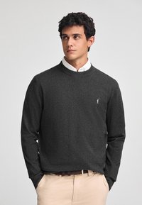 Maglione grigio scuro a maniche lunghe, con scollatura rotonda e un piccolo logo ricamato sul lato sinistro del petto. Indossato sopra una camicia bianca con colletto.