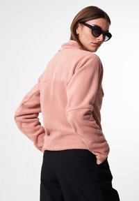 Helle rosa Fleecejacke mit hohem Kragen, ausgestattet mit einer Rückentasche und strukturiertem Stoff. Kombiniert mit schwarzen Hosen, die einen lässigen Schnitt zeigen.