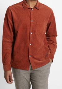 Camisa de corduroy de color óxido con collar tipo button-down, bolsillo frontal y botones blancos. Combinada con pantalones a cuadros con estampado.