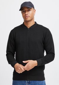 Sudadera con cremallera negra con diseño de textura de canalé, cuello redondo y puños ajustados. El modelo lleva una gorra oscura y jeans azules.