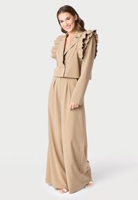 Smėlio spalvos pritaikytas kostiumas su trumpa švarku ir plištais pečiais bei plačiomis kelnėmis. Lygi tekstūra ir tvarkingos linijos sustiprina dizainą.