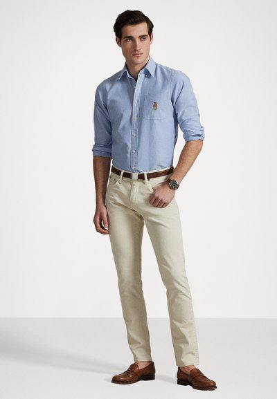 Chemise bleu clair à manches longues avec une poche poitrine et un logo brodé, associée à un pantalon beige slim-fit et des chaussures en cuir marron.