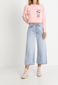 Ljust rosa kortärmad tröja med grafiska tryck, i kombination med ljusblå vida denimbyxor och beige högskurna sneakers.