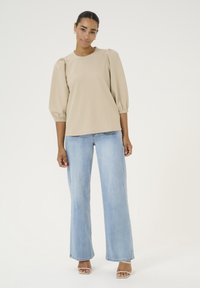 Beige puff-sleeve top with round neckline, paired with light blue wide-leg jeans and beige heels, on a plain white background.