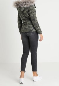 Puffjakke i camouflage mønster med en pelsforet hætte, tætsiddende design og en taljelængde, parret med tætsiddende mørke jeans og hvide sneakers.