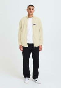Cremefarbene Fleecejacke mit hohem Kragen, seitlichen Taschen und schwarzem Logo-Patch, kombiniert mit einem weißen T-Shirt und schwarzen Hosen.