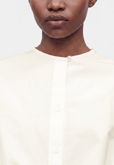Gros plan d'une personne portant une chemise crème boutonnée avec un col rond et une texture minimale sur la peau.