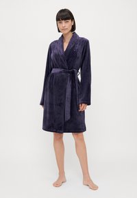 Lila badrock i mjuk, plyschig textil. Har långa ärmar, ett bälte i midjan och en scallop-mönstrad krage. Kjolen faller till mitten av låret.