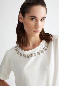 Maglietta in cotone bianco con scollo rotondo, caratterizzata da un decorativo ornamento dorato e nero attorno al colletto.