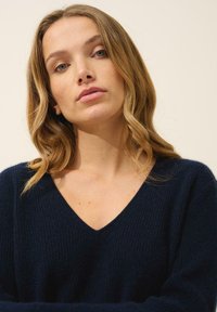 Granatowy sweter z dzianiny z głębokim dekoltem w kształcie litery V, z prążkowaną teksturą. Modelka ma falowane włosy i jest lekko nachylona do przodu.