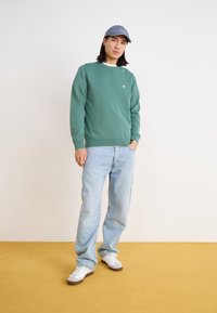 Green knitted sweater with ribbed cuffs and hem, paired with light blue denim jeans. White sneakers with grey accents. Model poses on a yellow floor.