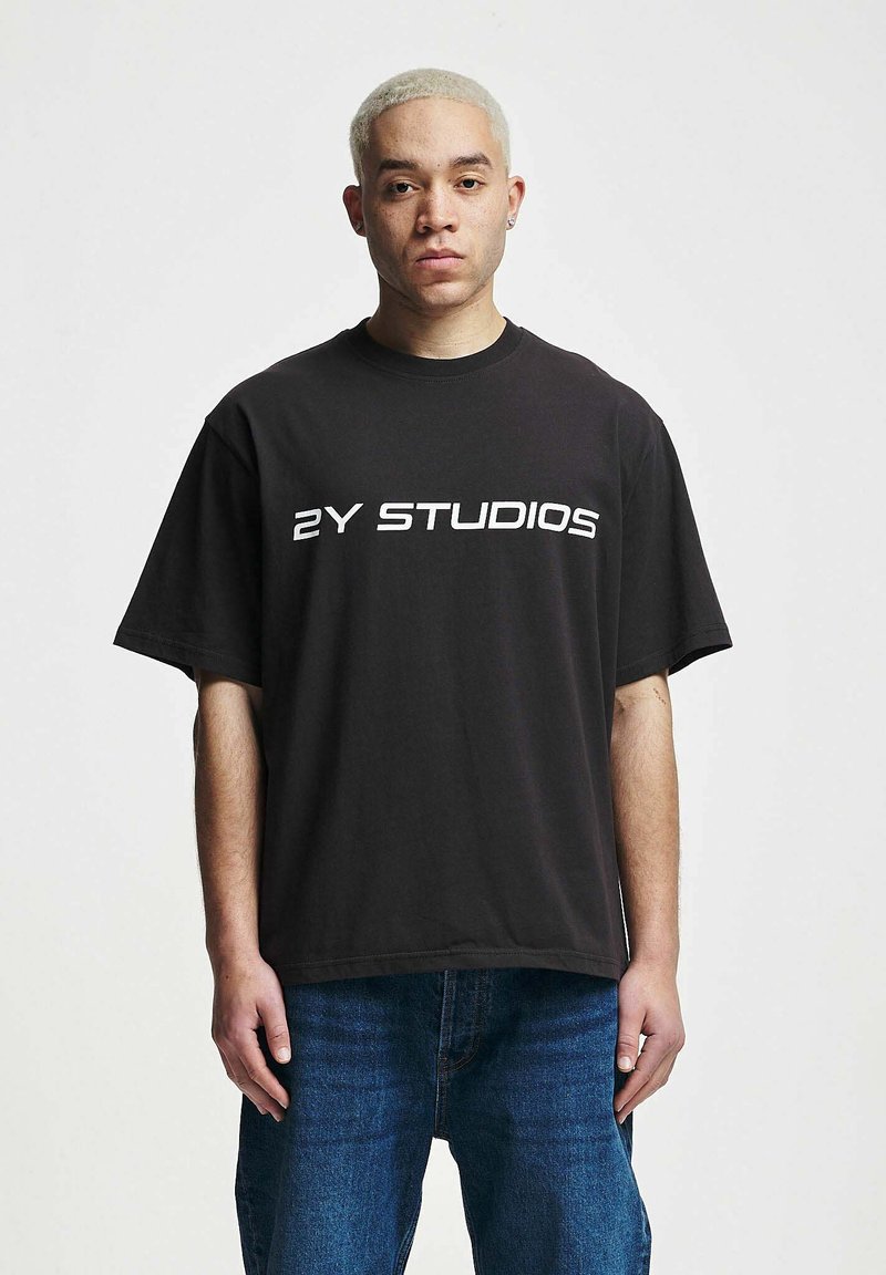 Musta puuvillainen t-paita lyhyillä hihoilla, jossa on rohkeaa valkoista tekstiä "2Y STUDIOS" rinnan kohdalla. Paritettu sinisten farkkujen kanssa.