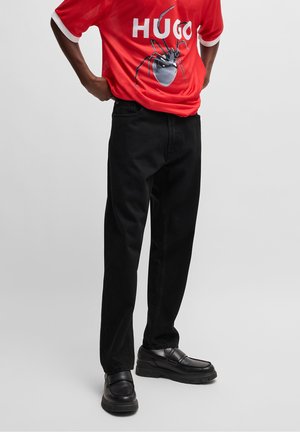 Personne portant un pantalon noir et des mocassins noirs avec un T-shirt rouge présentant un grand graphisme d'araignée et un texte blanc "HUGO".