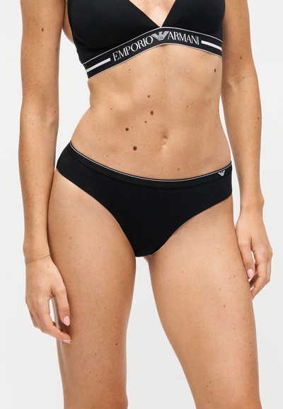 Femme portant un soutien-gorge noir de la marque Emporio Armani et une lingerie assortie sur un fond clair uni.