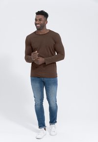 Braunes Langarmshirt mit Rundhalsausschnitt, kombiniert mit hellblauen Jeans und weißen Sneakers. Glatter Stoff und körpernahe Passform.