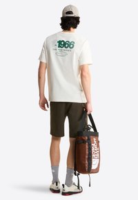 Hombre con gorra beige, camiseta blanca The North Face 1966, pantalones cortos oscuros, calcetines grises, zapatillas blancas, sujetando una bolsa de viaje marrón de The North Face a un lado.