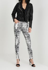 Femme portant une veste en cuir noir, un haut noir, un pantalon à motifs noir et blanc, et des sandales à talons hauts noires, se tenant avec une main dans la poche.