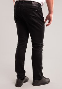 Fekete farmert és fekete tornacipőt viselő férfi, aki egy egyszerű világosszürke háttér előtt áll, hátulnézetben.