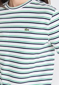 Randig t-shirt i bomull, med smala marinblå och gröna ränder på en vit botten. Litet broderat grönt krokodillogo på bröstet.