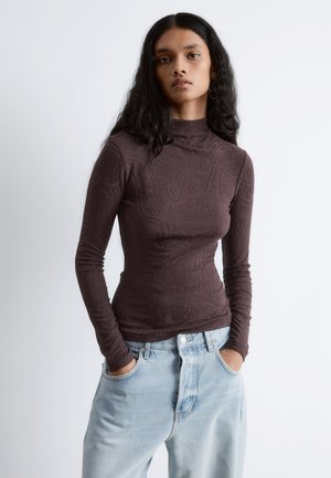 Long-sleeve, high-neck top in dark brown textured fabric with paisley pattern. Paired with light blue high-waisted denim jeans.