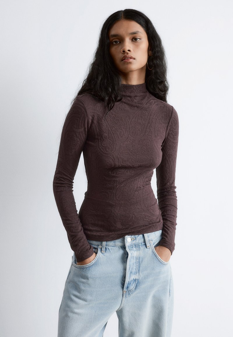 Langärmliges Oberteil mit hohem Kragen aus dunkeltbraunem, strukturiertem Stoff mit Paisleymuster. Kombiniert mit hellblauen, hoch taillierten Jeans.