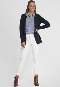 Cardigan en maille navy sur une chemise rayée bleue et blanche, associé à un jeans skinny blanc et des bottines brunes.