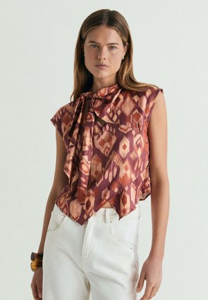 Mujer con blusa sin mangas, estampada en burdeos y naranja con escote anudado, combinada con pantalones blancos de talle alto y pulseras marrones.